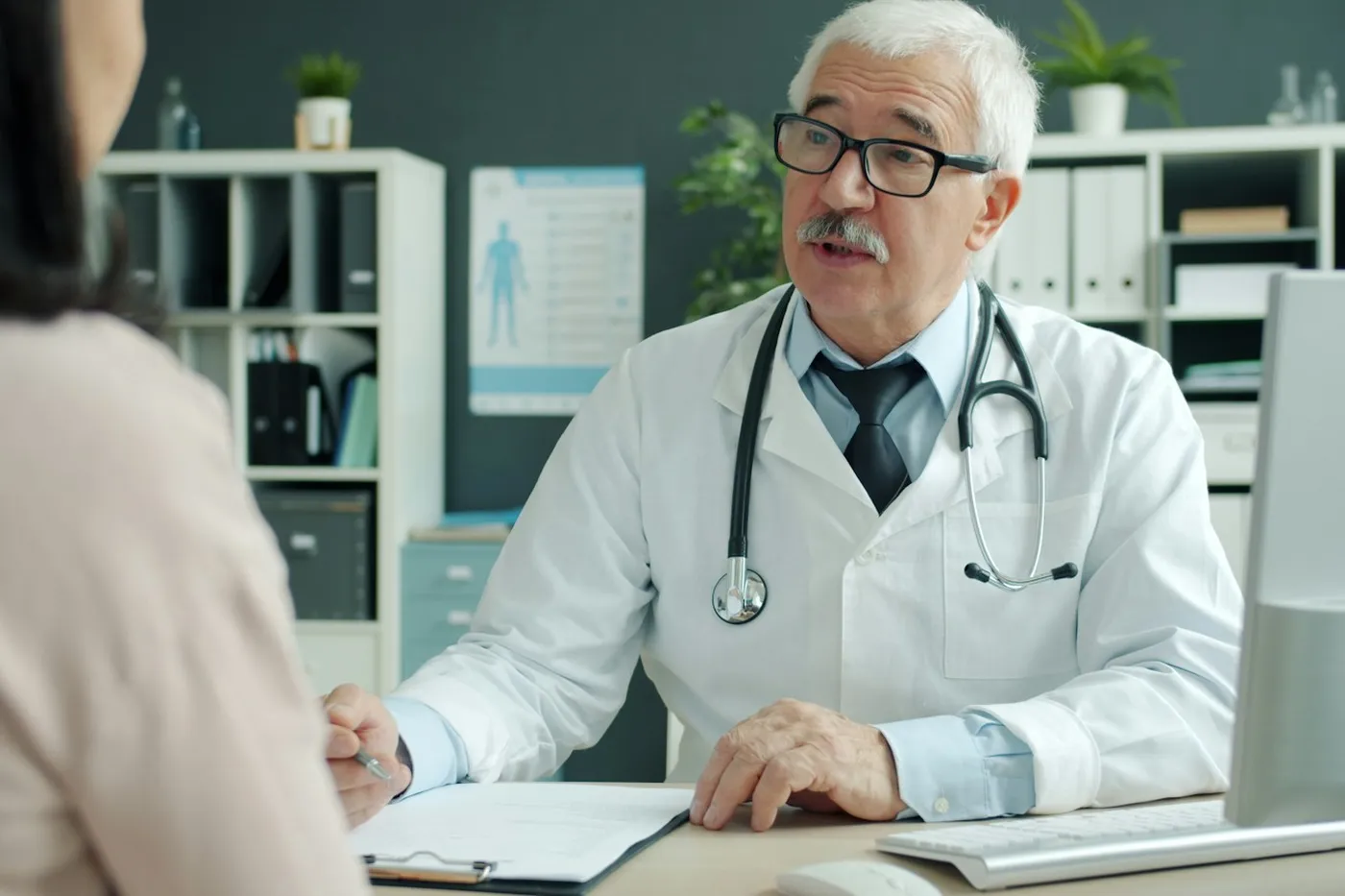 Médico de jaleco branco e estetoscópio conversando com uma paciente, sentados frente a frente em uma mesa de consultório