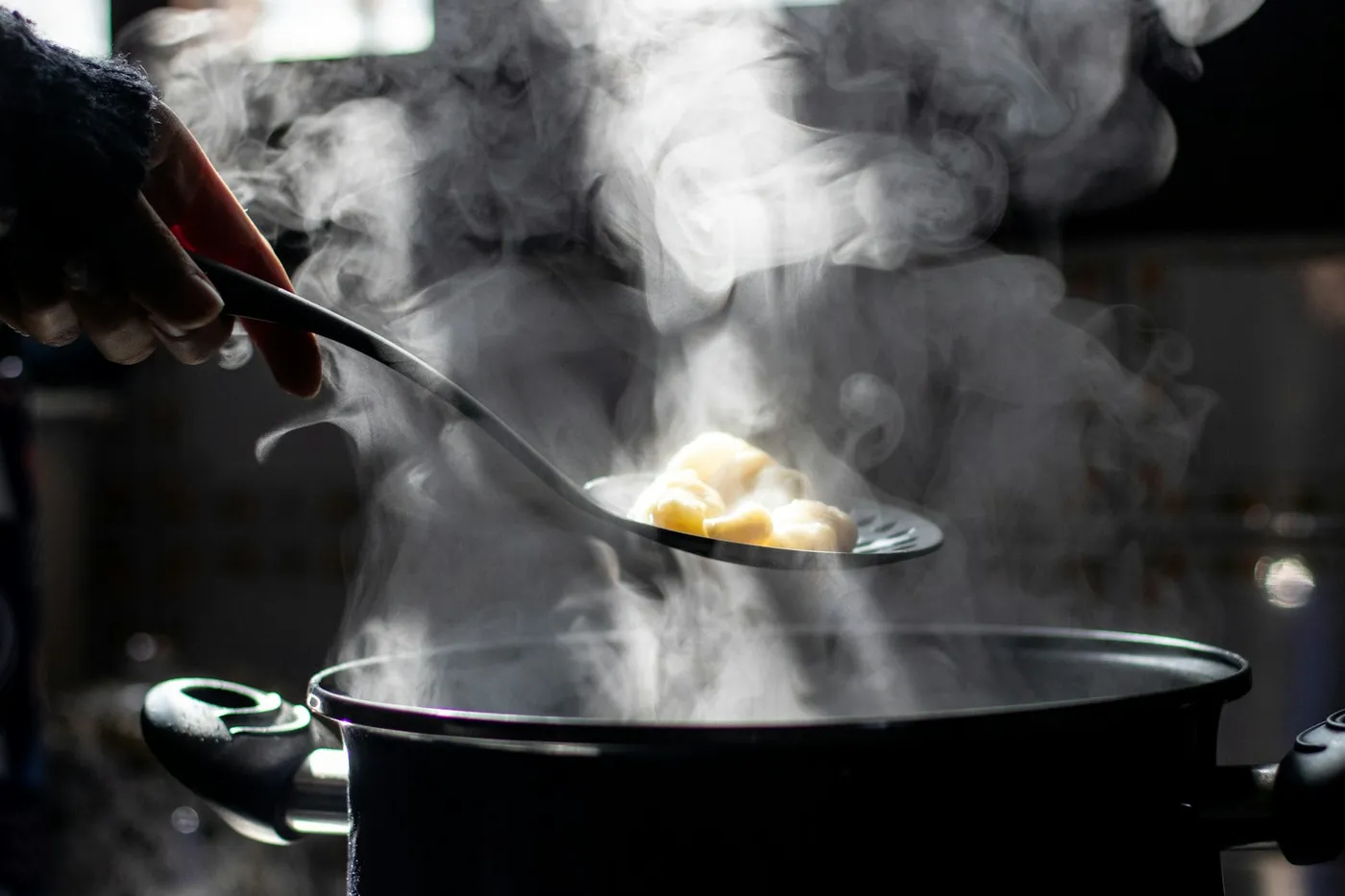 Mão segurando uma escumadeira com nhoque sobre uma panela escura que solta muito vapor na cozinha
