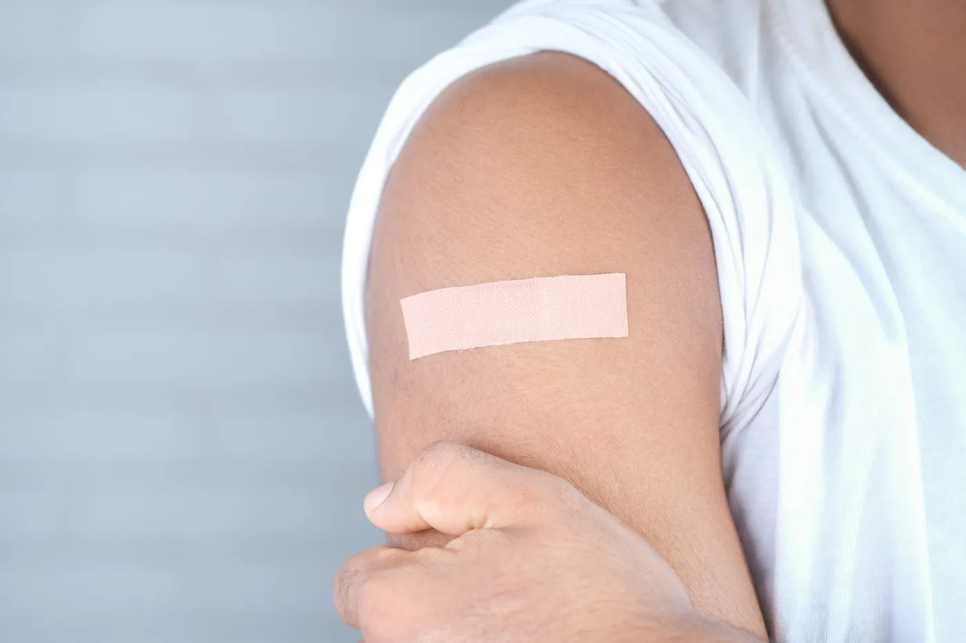 Braço de um jovem de camiseta branca com um curativo bege na região do ombro, logo após receber uma vacina