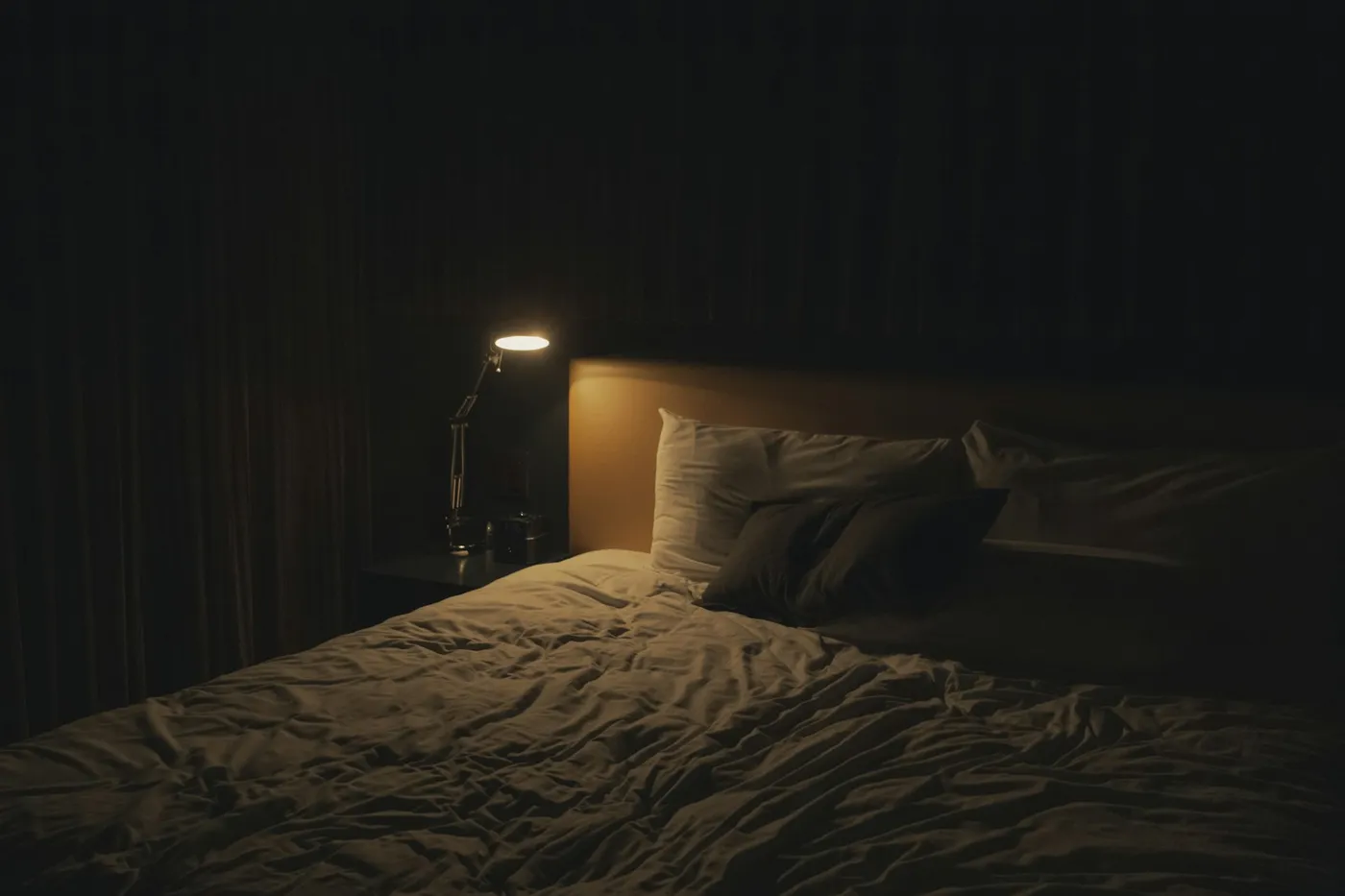 Cama de casal desarrumada num quarto escuro, com travesseiros brancos e um abajur de braço articulado aceso sobre o criado-mudo