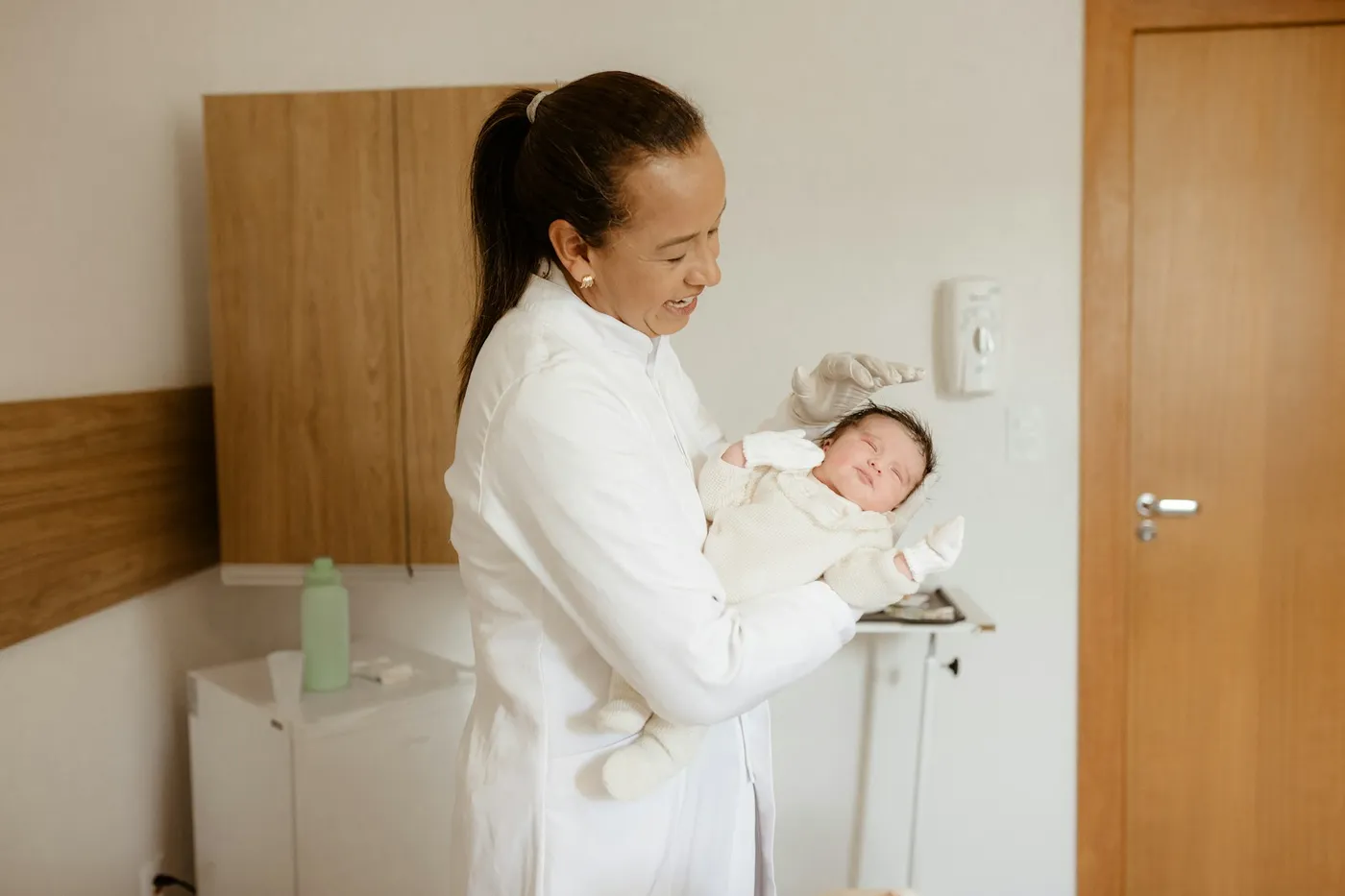 Profissional de saúde de jaleco branco e cabelo preso sorri para um recém-nascido adormecido, de macacão branco, que ela segura no colo numa sala de atendimento com armário de madeira