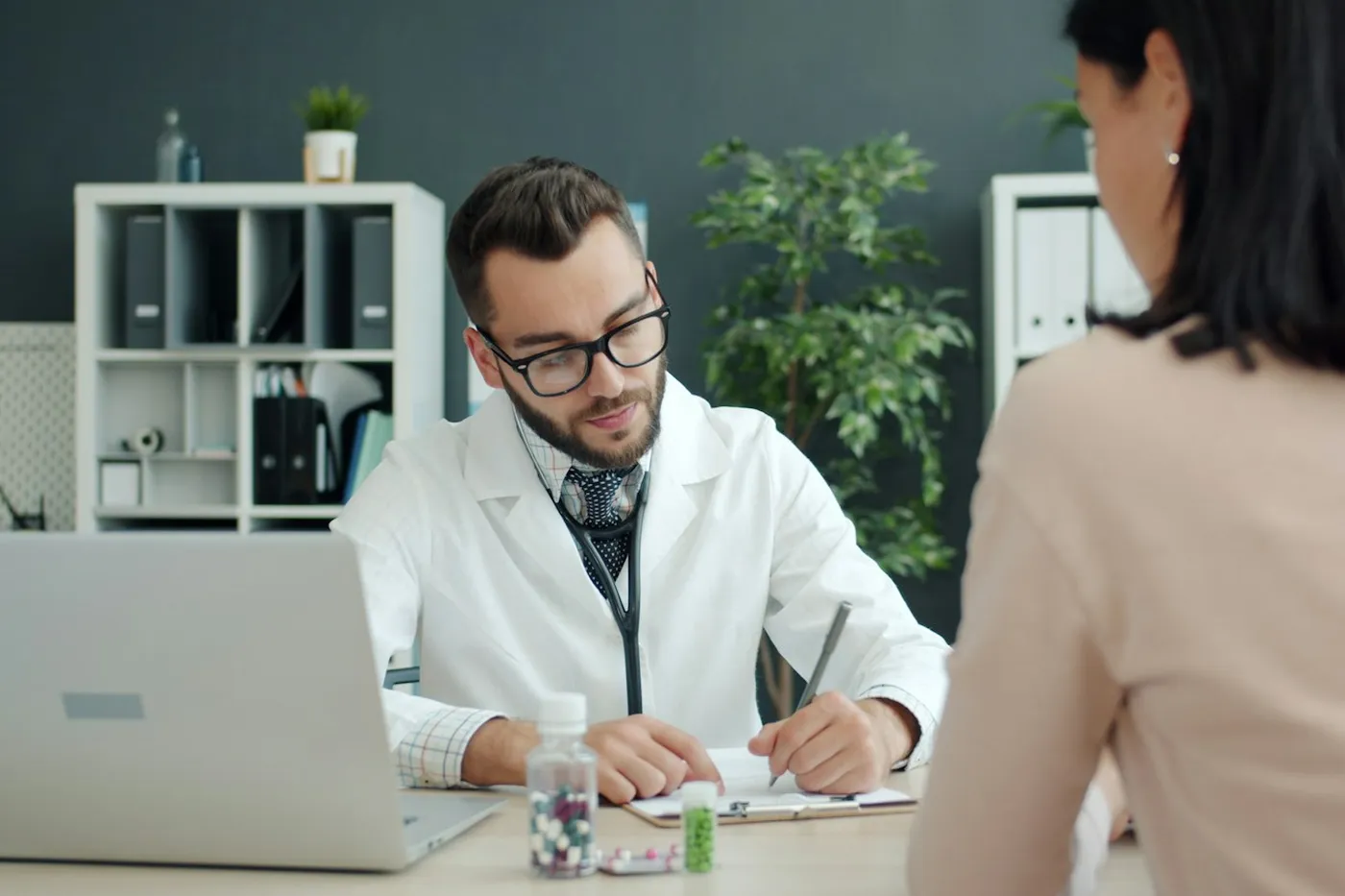 Médico de jaleco branco e óculos fazendo anotações à mesa enquanto conversa com uma paciente de cabelo escuro, em consultório com laptop e plantas