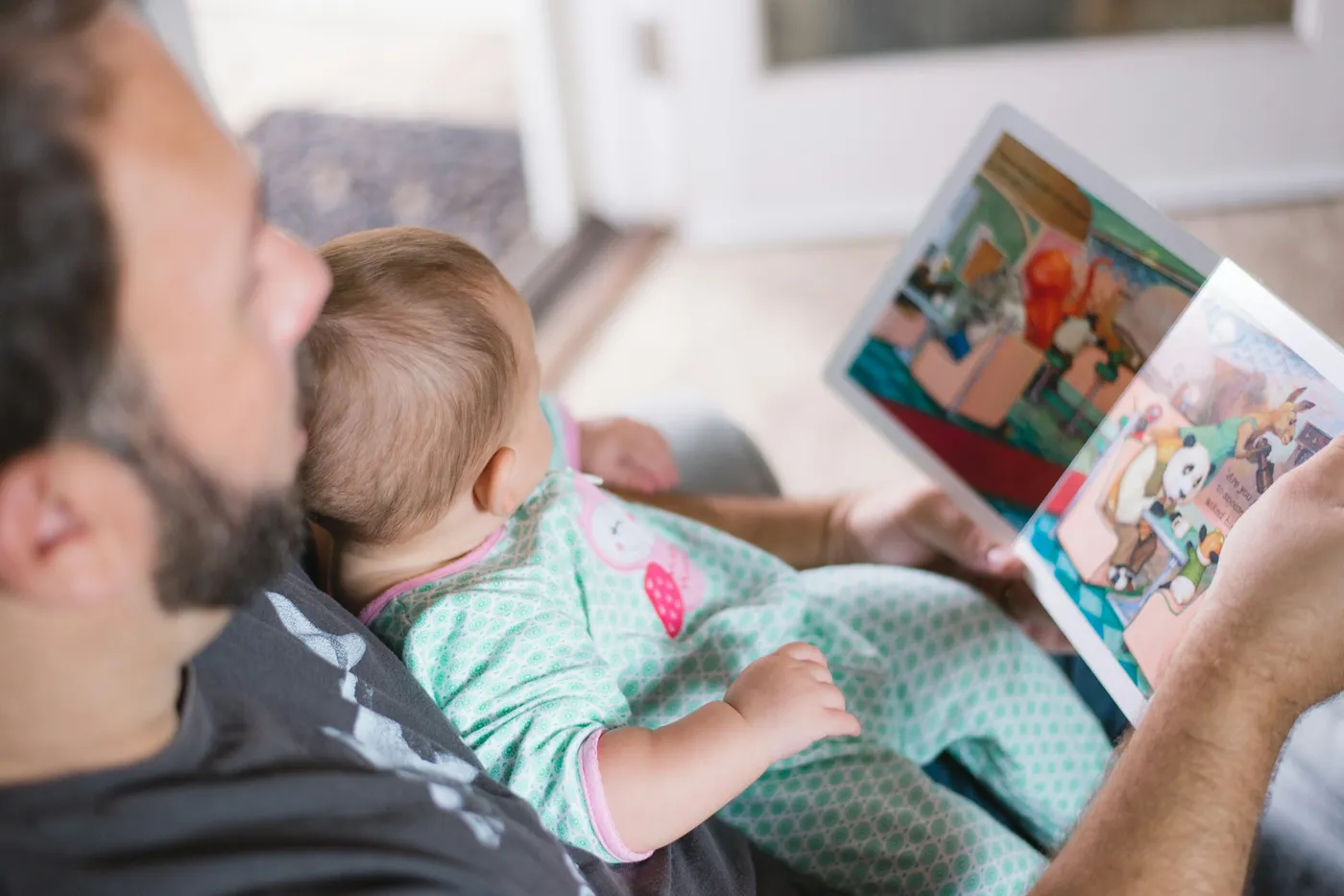 Homem de barba e camiseta cinza com um bebê de macacão verde estampado no colo, segurando aberto um livro infantil ilustrado com bichos