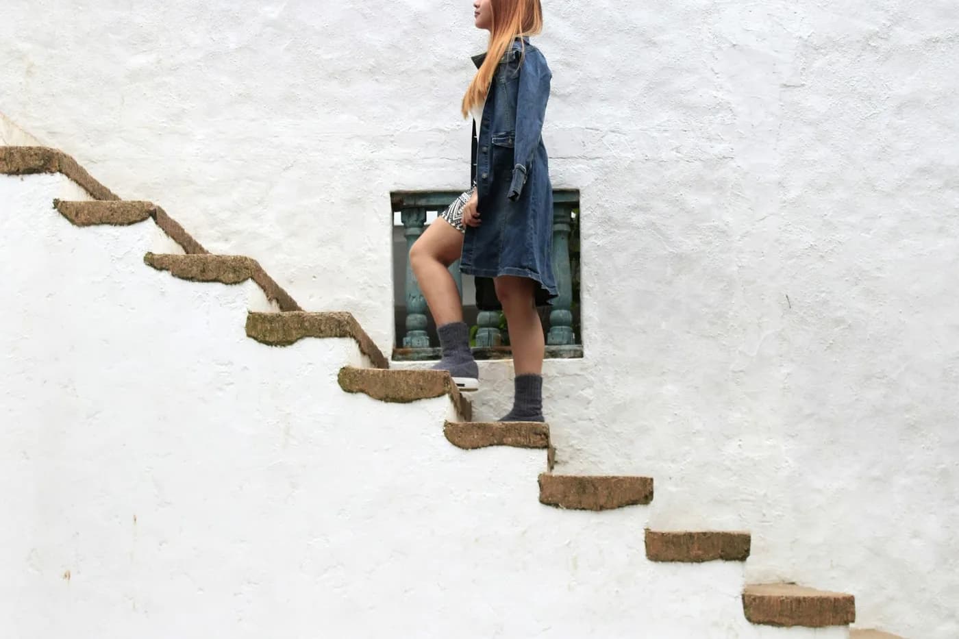 Mulher de jaqueta jeans subindo uma escada de degraus de pedra encostada em um muro branco