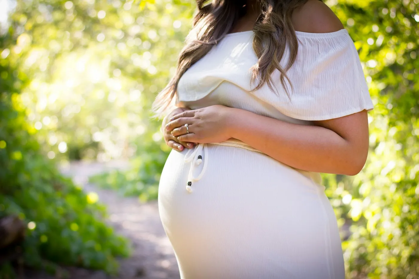 Gestante de vestido branco em uma trilha arborizada, com as duas mãos apoiadas na barriga