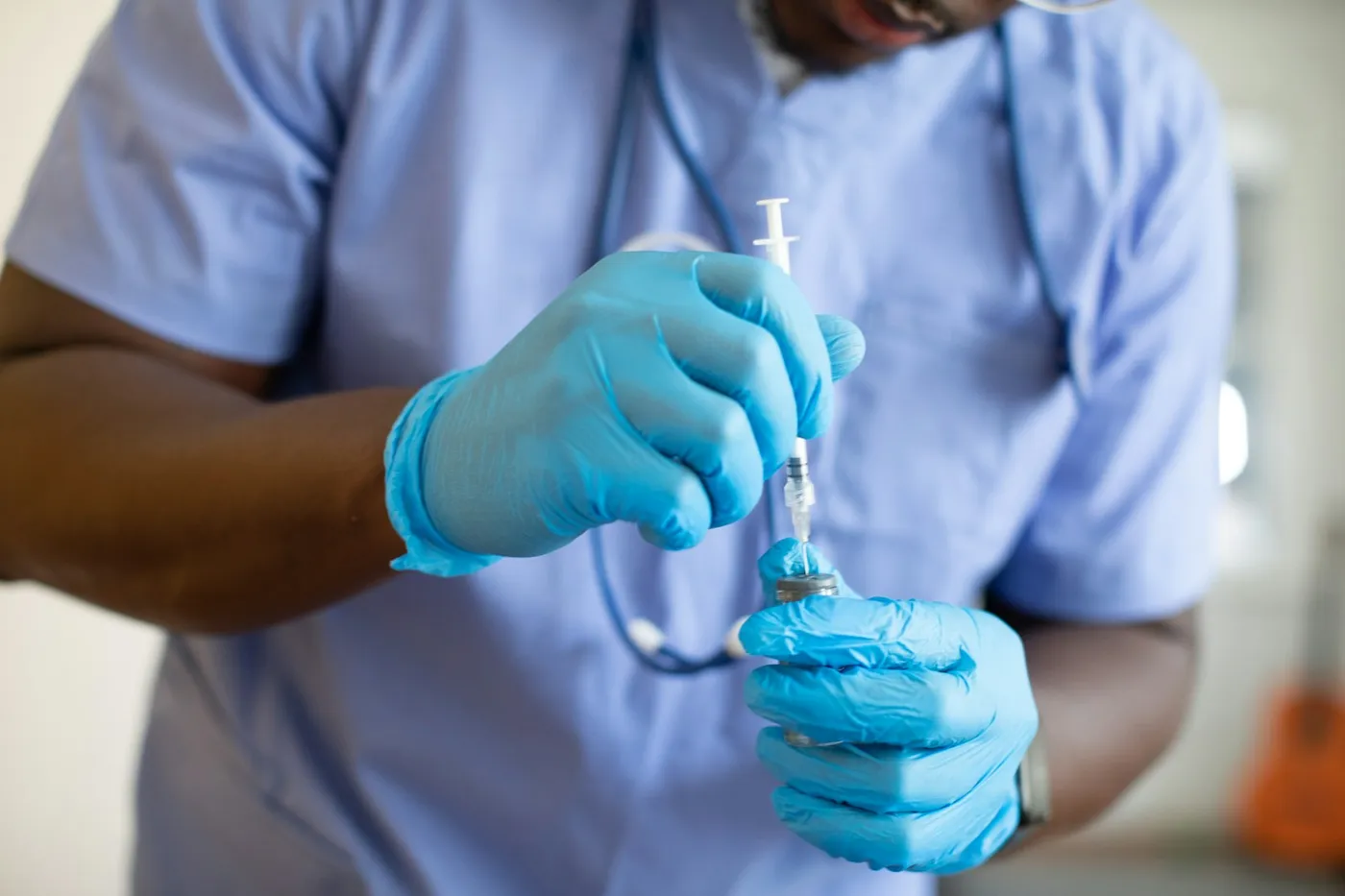 Profissional de saúde de jaleco azul e luvas azuis aspirando o conteúdo de um frasco com uma seringa