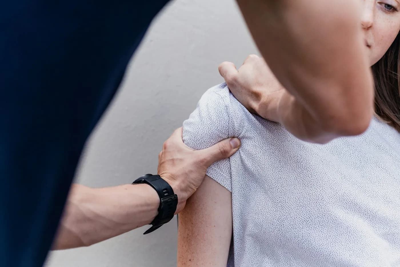 Profissional de saúde segura o ombro de uma mulher de camiseta branca durante uma avaliação, contra uma parede clara