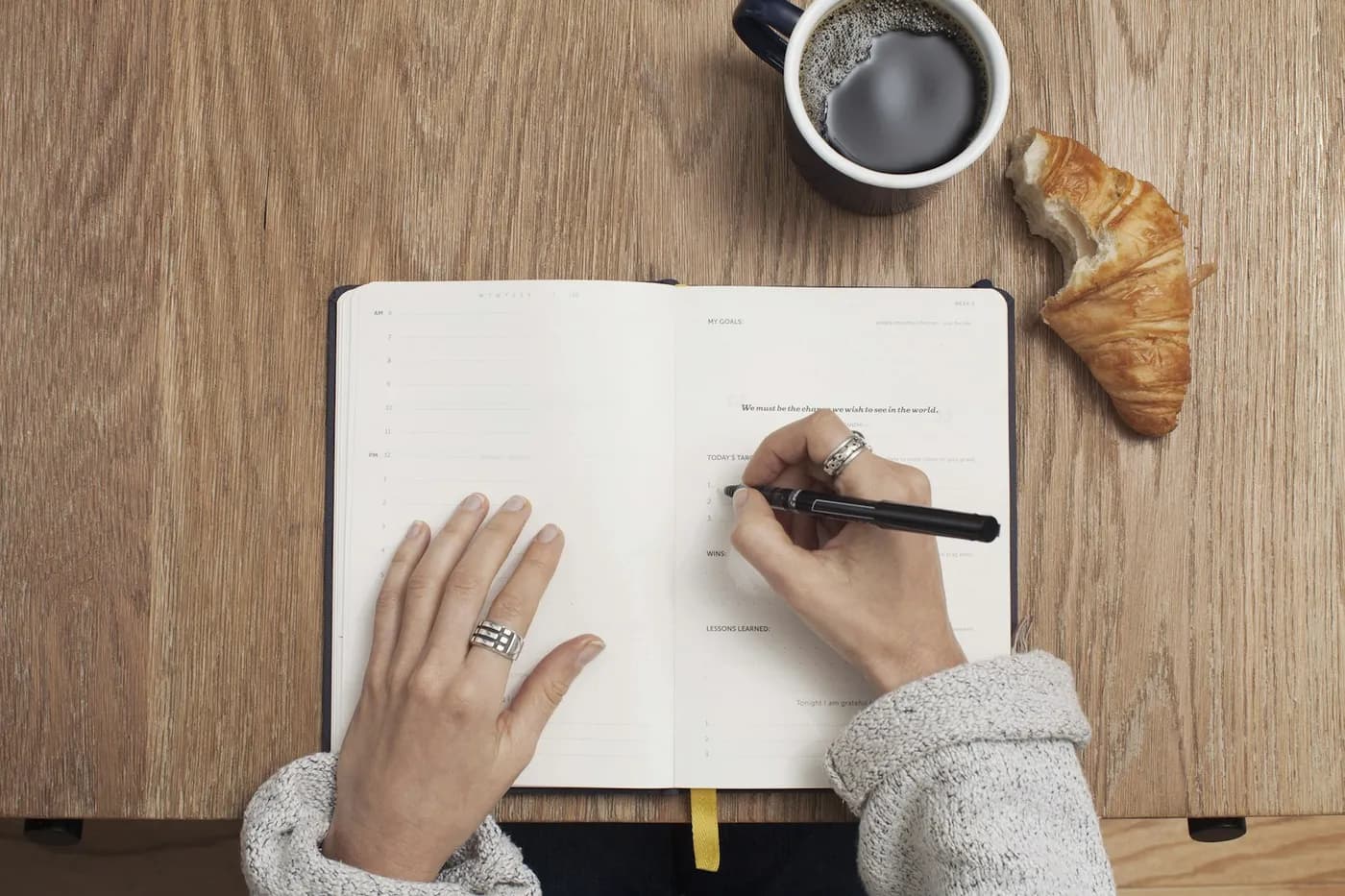 Mãos de uma pessoa de suéter cinza escrevendo em um caderno aberto sobre uma mesa de madeira, ao lado de uma xícara de café e de um croissant