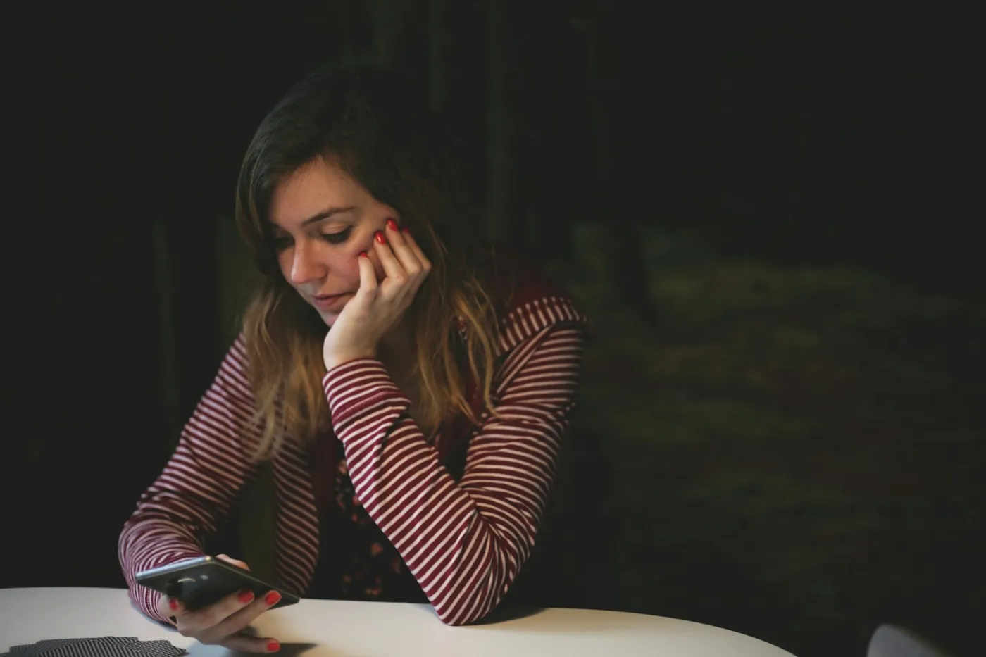 Mulher de blusa listrada sentada à mesa, com a mão no rosto, olhando para o celular em um ambiente escuro