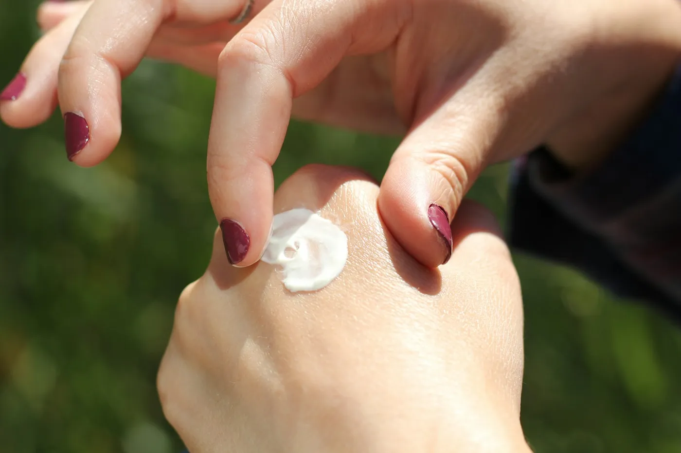 Dedos de unhas vinho espalhando uma porção de creme branco no dorso da mão, ao ar livre, com fundo verde desfocado